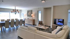 Uma sala de estar com sofá, mesa de jantar, cadeiras, um lustre, televisão e um relógio na parede.