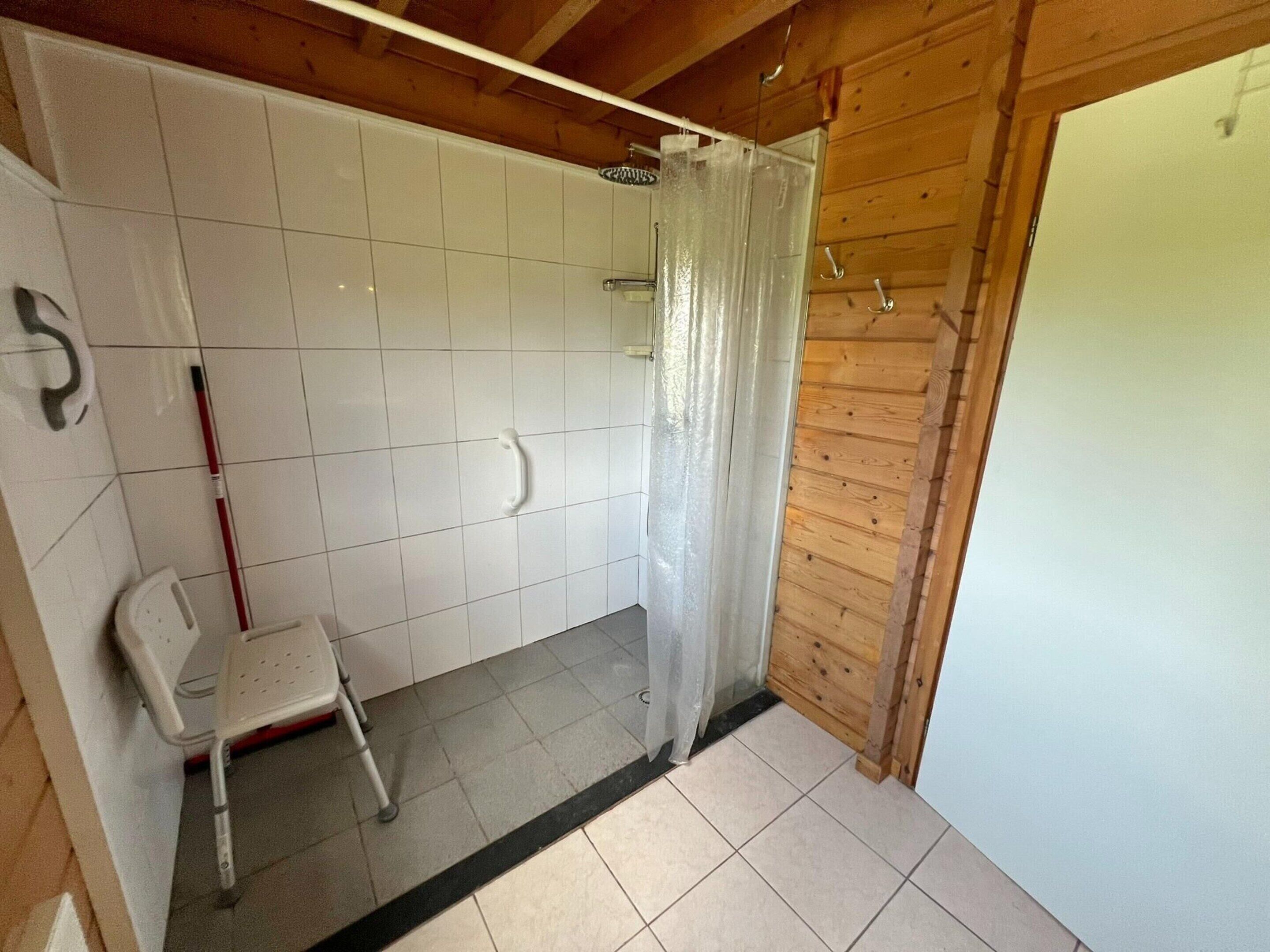 A bathroom with white tiled walls, a shower with a glass door, a white plastic chair, and a wooden ceiling.