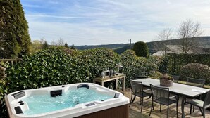 A hot tub with a wooden bench, surrounded by a table and chairs, set against a backdrop of a hedge and a view of hills. - Luxurious Holiday Home With Sauna in Fauvillers (Fauvillers)