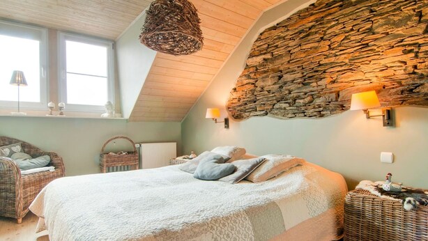 A cozy bedroom with a sloped ceiling, a stone wall, a large bed with pillows, a wicker chair, and a hanging light fixture. - Luxurious Holiday Home With Sauna in Fauvillers (Fauvillers)