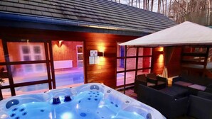 A hot tub with multiple jets, a wooden deck, outdoor seating, and a view of a building with a glass door. - Holiday Home in Stoumont With Private Sauna (Stoumont)