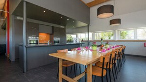 A modern dining area with a long wooden table set for multiple guests, a kitchen area with stainless steel appliances, and large windows providing natural light. - Comfy Home with Sauna Located near Forest (Waimes)