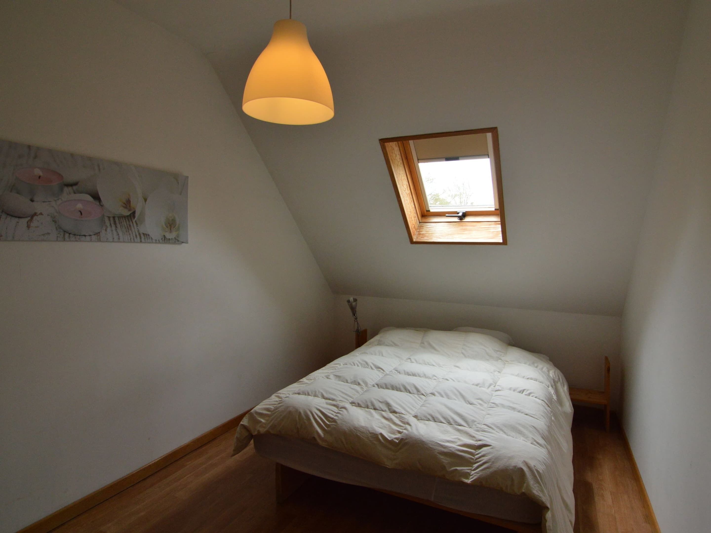 Une chambre avec un plafond en pente, un lit simple, une petite fenêtre et un luminaire suspendu.