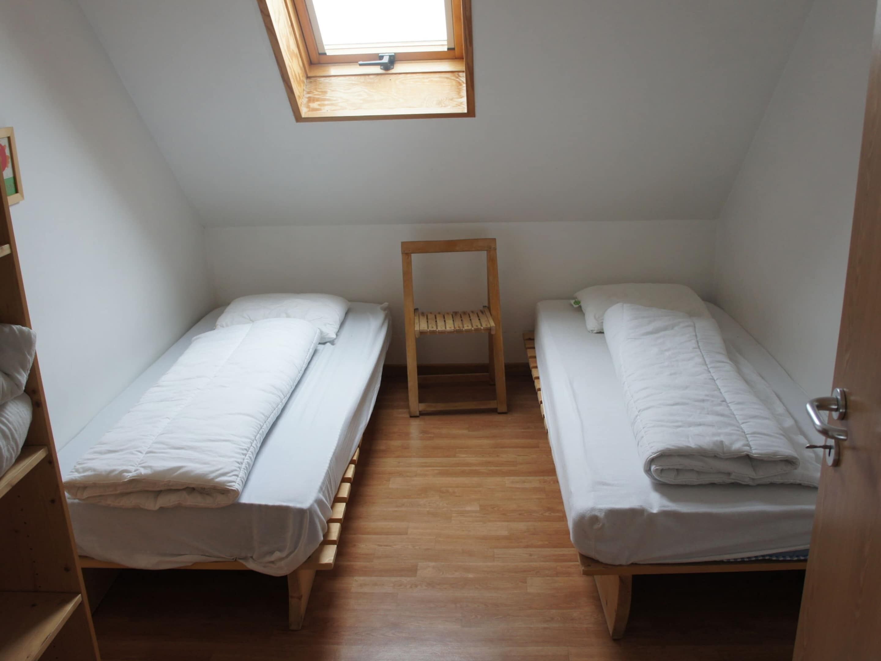 Un alojamiento pequeño y de una sola habitación con dos camas individuales, una silla de madera y un tragaluz.