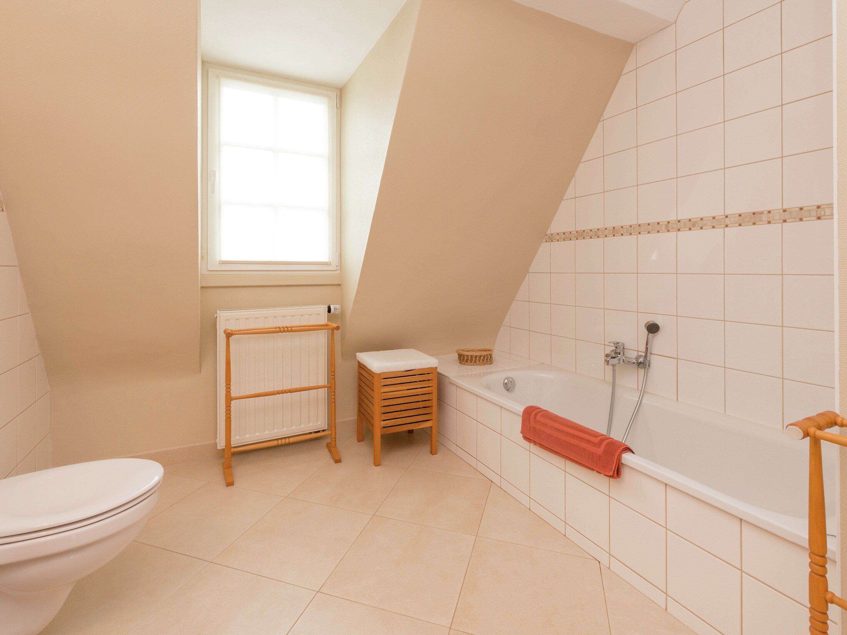 A bathroom with a toilet, a wooden drying rack, a small wooden stool, a bathtub with a showerhead, and a window providing natural light.