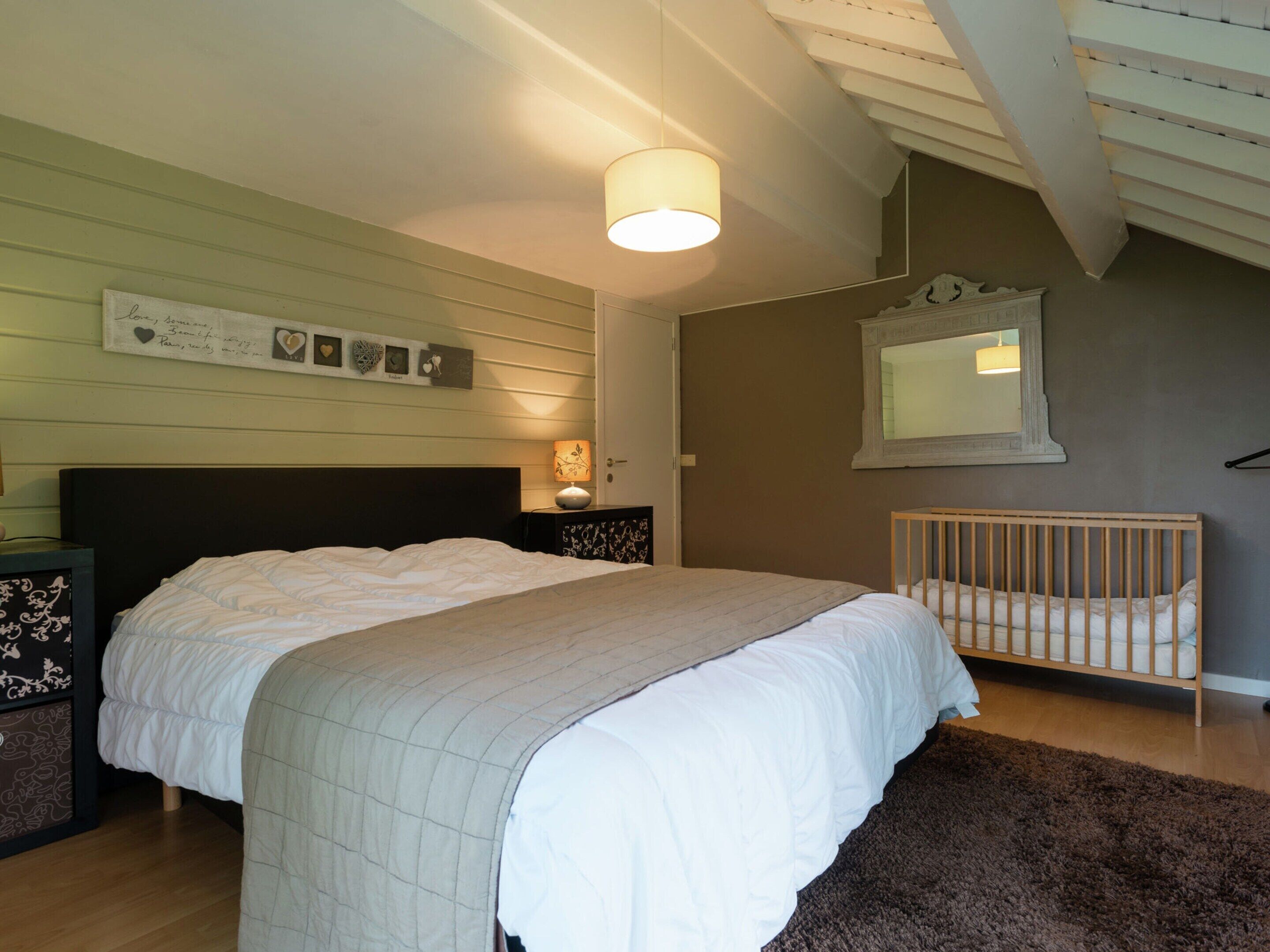 Un dormitorio con una cama grande, una mesita de noche con lámpara, una cuna y un espejo instalado en la pared.