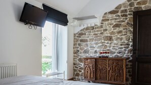 Een slaapkamer met een groot bed, een stenen muur, een houten kast met gravures, een aan de muur bevestigde televisie en een raam met uitzicht op groen.