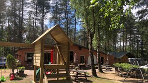 Une structure de jeux en bois située dans une zone boisée, avec des tables de pique-nique et un bâtiment en briques en arrière-plan.