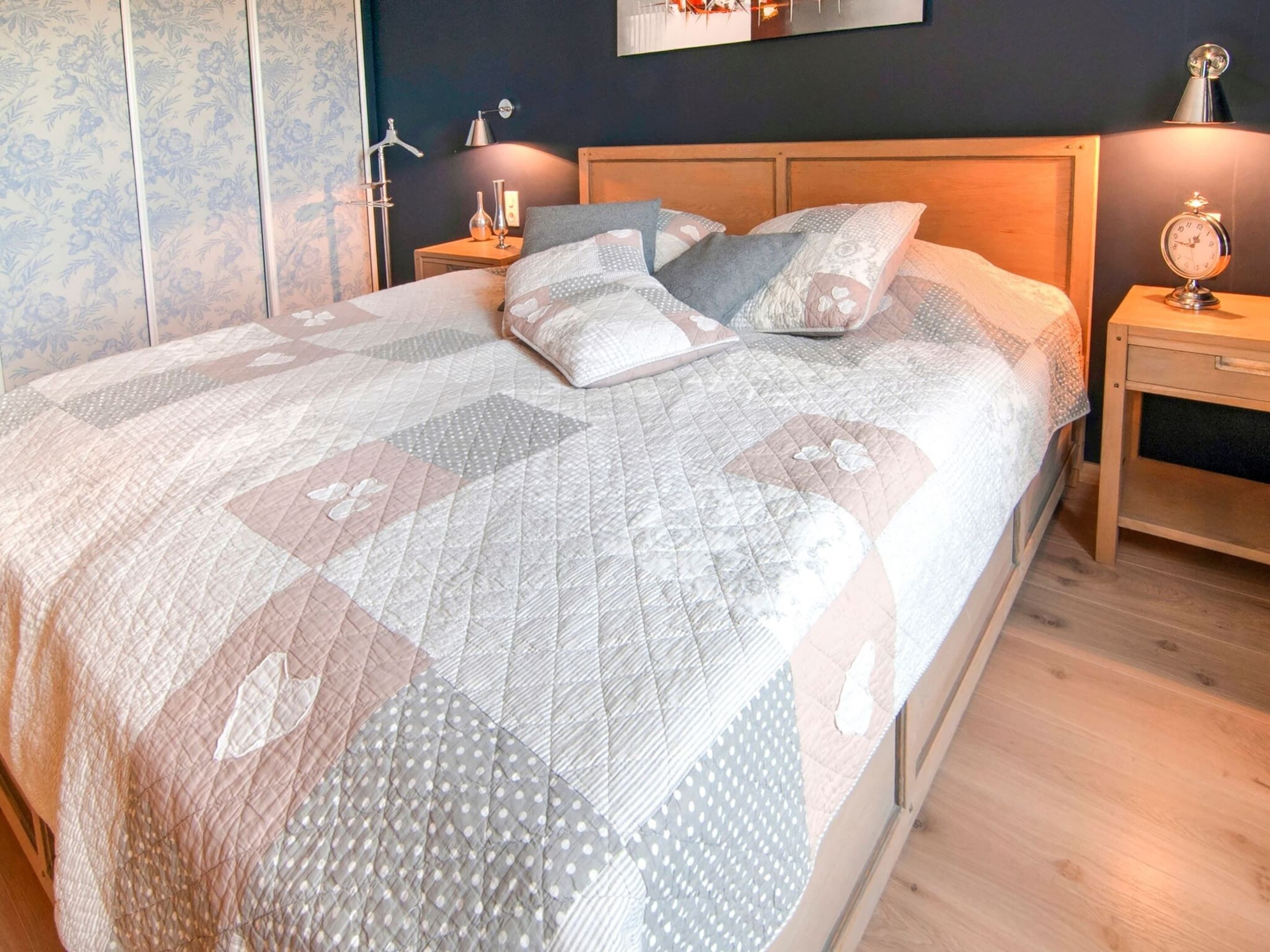 Een net opgemaakt bed met een sprei, een nachtkastje met een lamp en een wandklok.
