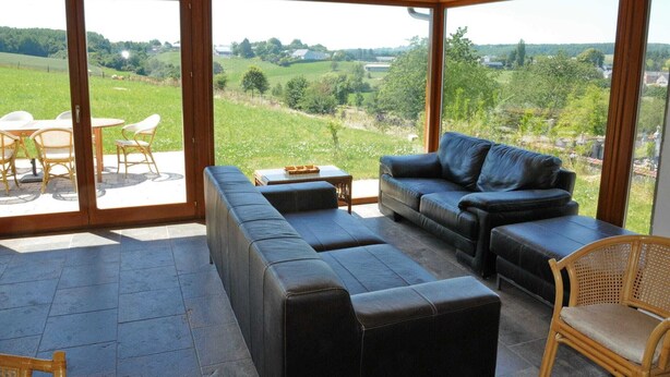 A spacious room with a large sliding glass door leading to an outdoor area. Inside, there is a black leather sofa, a wooden coffee table, and wicker chairs. - Stone House in Durbuy With Sauna & Garden Views (Somme-Leuze)