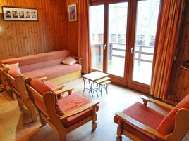 Una habitación acogedora con detalles de madera, un sofá, dos sillas de madera, una mesita y un ventanal corredero que da a un espacio exterior.