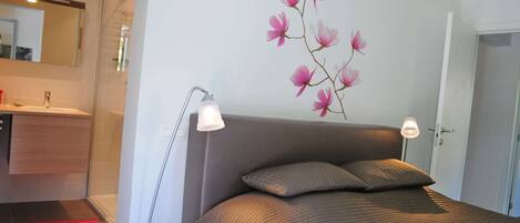 A bedroom with a bed, bedside lamps, a wall-mounted mirror, and a bathroom visible through an open door.