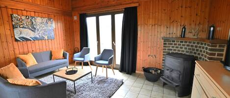 Ett mysigt vardagsrum med träpanel på väggarna, en soffa, två fåtöljer, ett soffbord och en öppen spis.