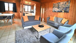 L’intérieur d’un chalet accueillant comprend un coin repas, deux canapés avec des coussins, une table basse et une cuisine visible à travers une porte ouverte.