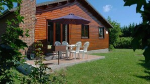Une cabane en bois avec une cheminée en briques, une terrasse avec des chaises et une table blanches, et un espace extérieur couvert.