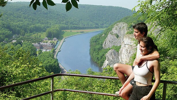 A couple sitting on a railing overlooking a river, with a lush green landscape and a castle in the distance. - Nicely Furnished Bungalow with Fireplace near Maas (Hastiere)