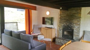 A cozy living room with a grey sofa, a small round table, a wooden cabinet, and a stone fireplace. - Nicely Furnished Bungalow with Fireplace near Maas (Hastiere)