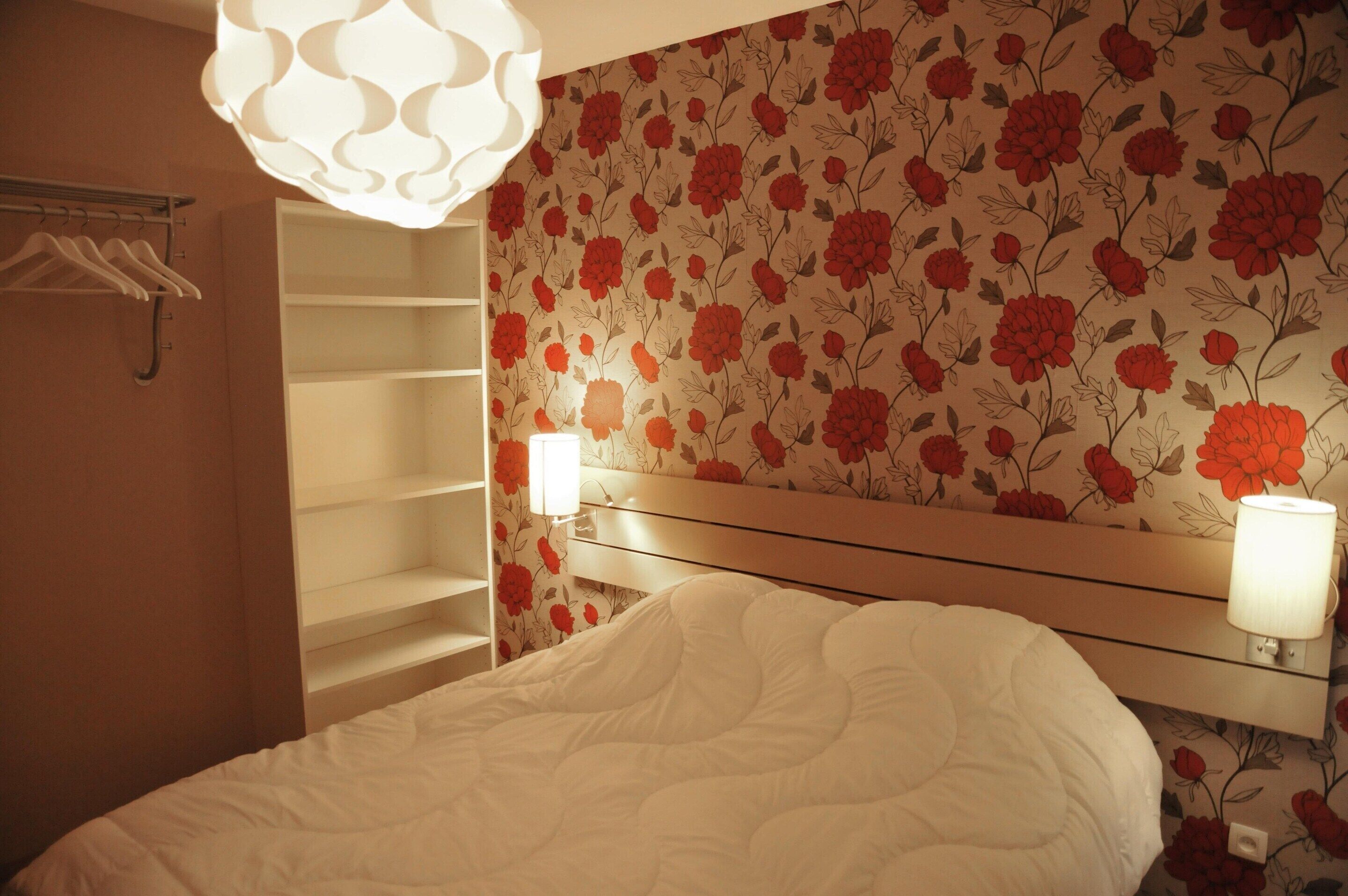 Ein Schlafzimmer mit einem Bett, einem Nachttisch mit Lampe, einem Kleiderschrank und floralem Wandpapier.