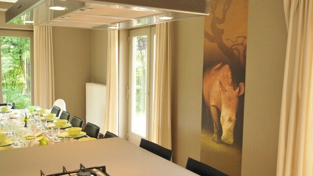 A dining area with a long table set for a meal, a gas stove, and a large window with curtains. - Holiday Home in Marche-en-famenne Near Train (Marche-en-Famenne)