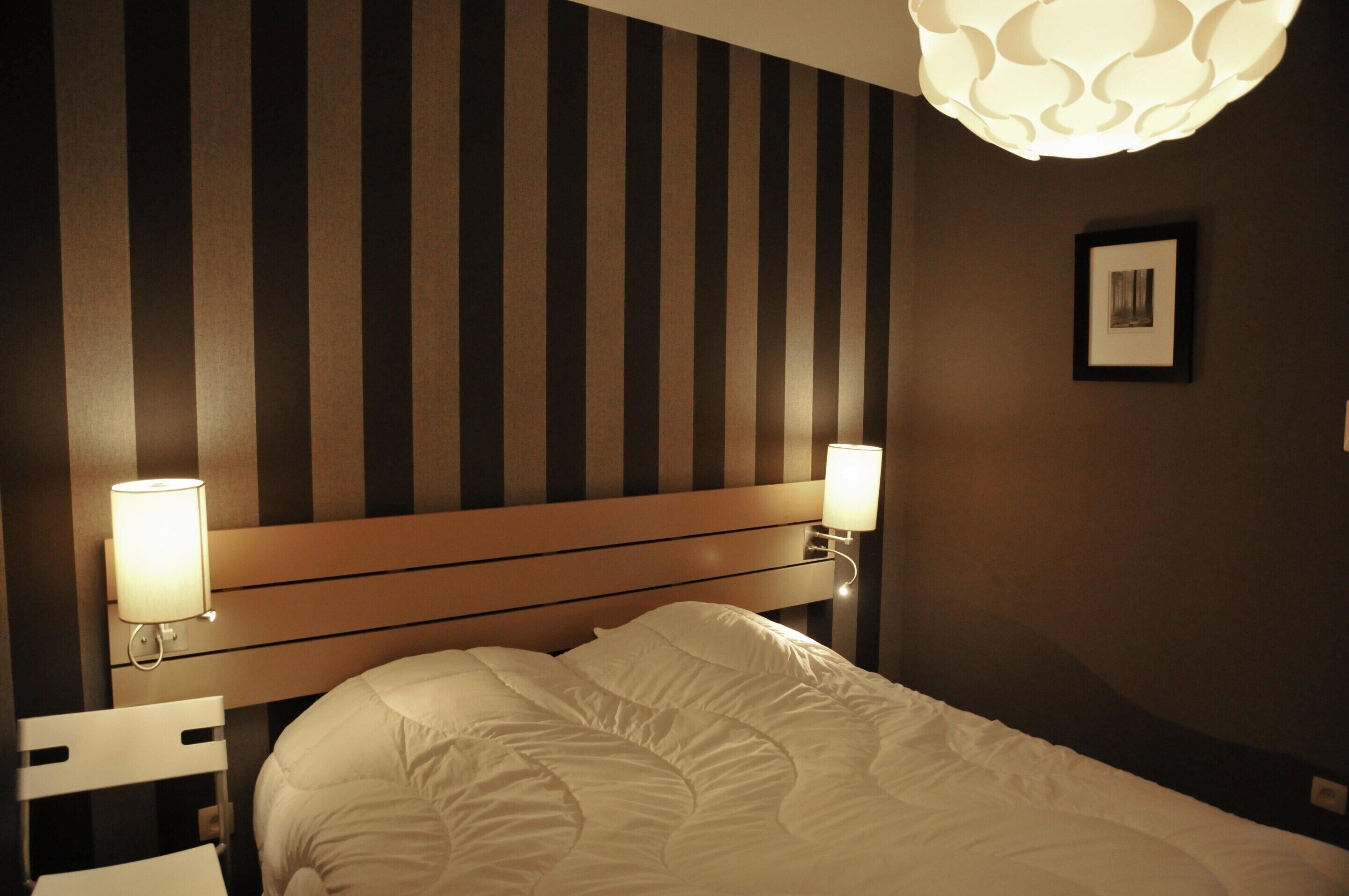 A hotel room with a striped wall, a wooden headboard, a bed with white bedding, two bedside lamps, and a framed picture on the wall.