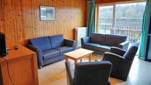 A room with wooden panel walls, a blue sofa, a wooden table, and a view of a lake through large windows. - Holiday Home in Vielsalm With Lake View (Vielsalm)