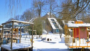 A snow-covered playground with climbing structures and a slide, surrounded by bare trees and a building with a sloped roof. - Holiday Home in Vielsalm With Lake View (Vielsalm)