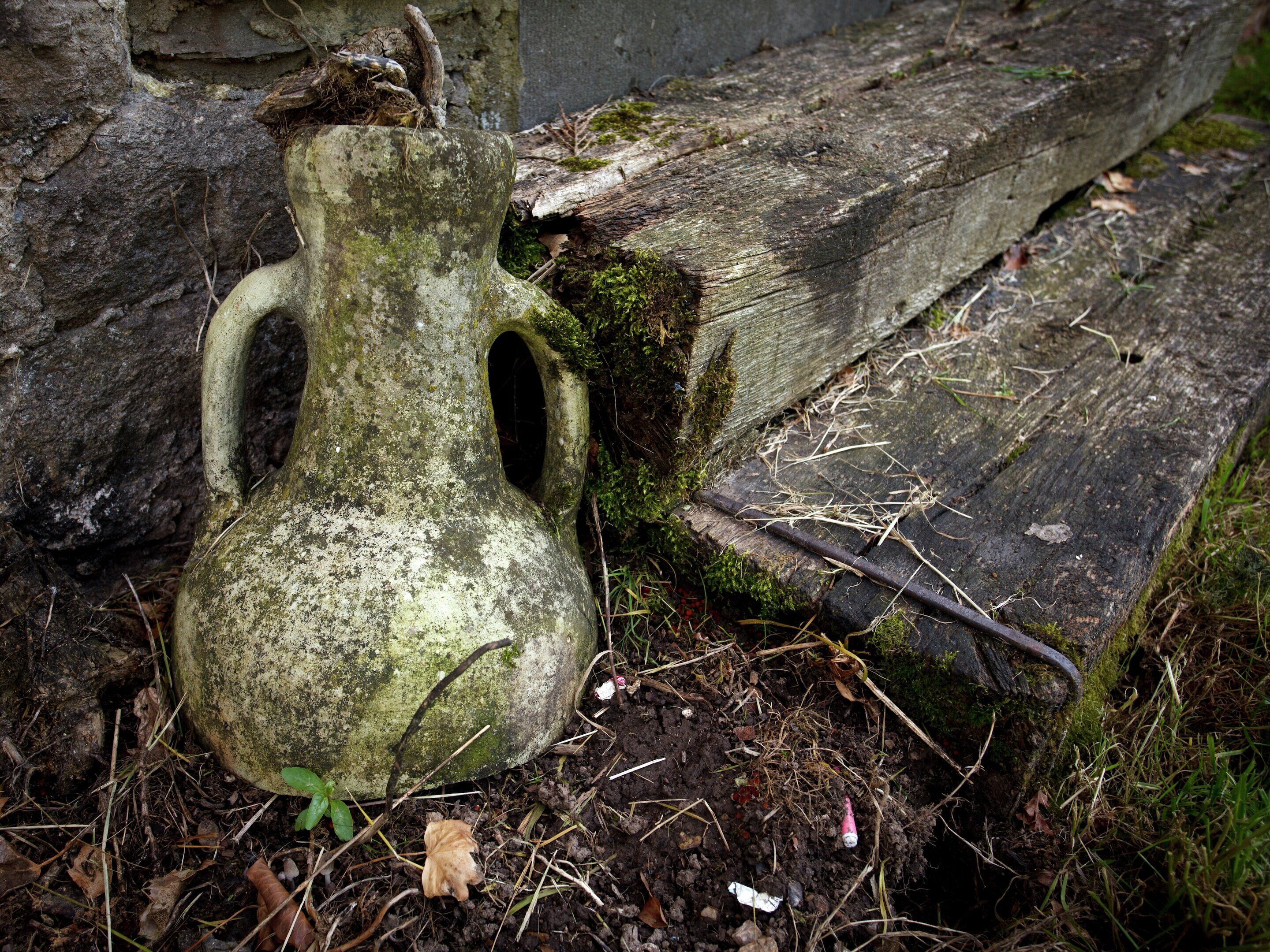 A weathered stone vase with a handle, surrounded by moss and debris.