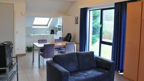 Una sala de estar moderna con un sofá, una mesa de comedor con sillas, una cocina al fondo y un amplio ventanal con cortinas.