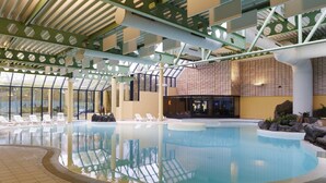 Una piscina interior con agua cristalina, rodeada de sillones de descanso y un diseño arquitectónico moderno con elementos estructurales verdes y ventanas amplias.