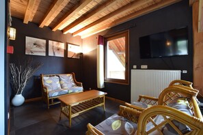 A room with a wooden ceiling, a sofa, a table, wicker chairs, a TV, and artwork on the wall. - Rendeux Retreat With Sauna (Rendeux)