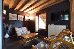 A room with a wooden ceiling, a sofa, a table, wicker chairs, a TV, and artwork on the wall.