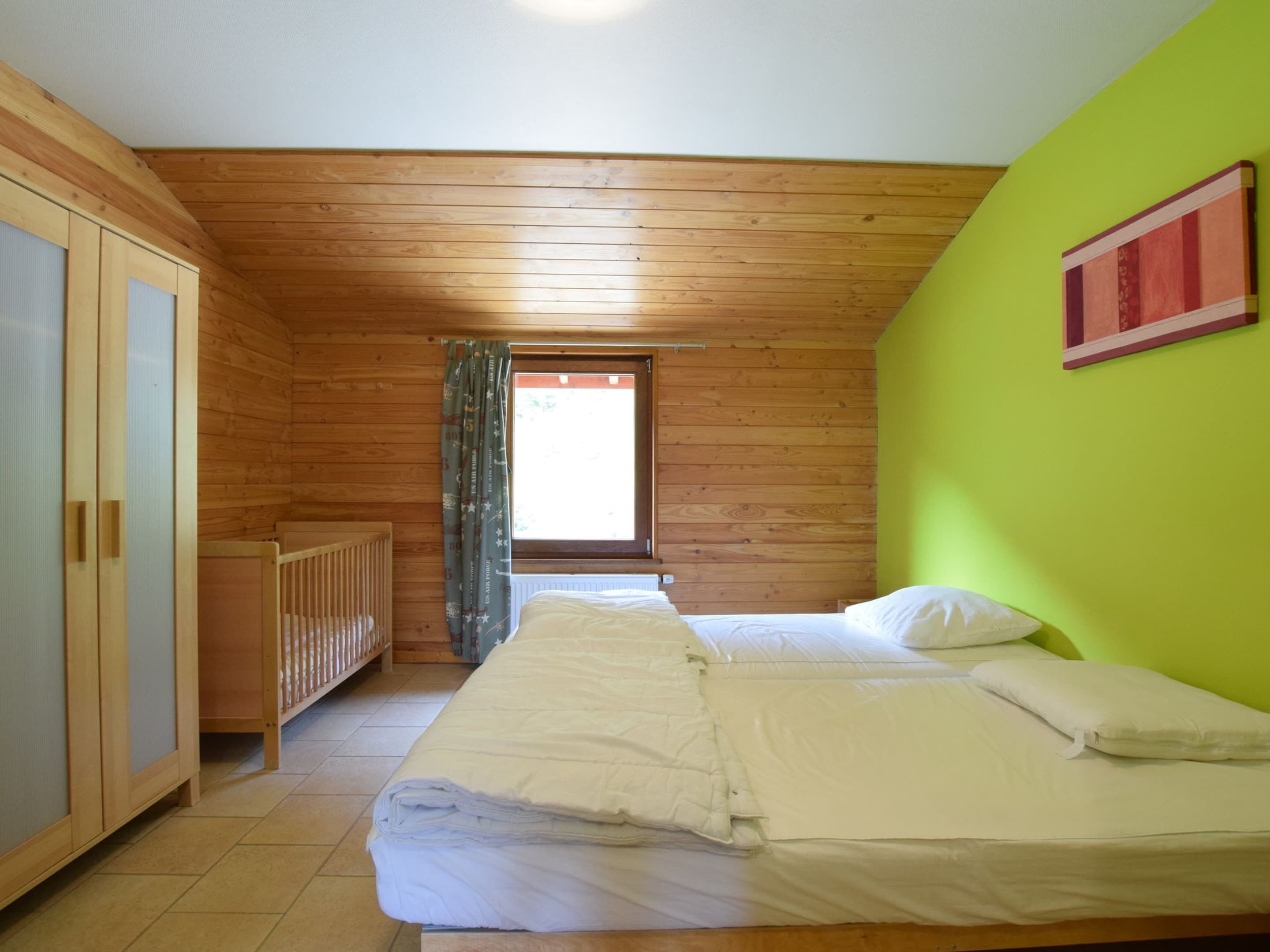 A bedroom with a wooden ceiling, a bed with white bedding, a crib, and a green wall.