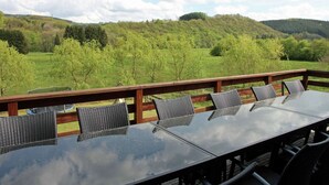 Una terraza con una mesa de vidrio y cuatro sillas, con vistas a un paisaje verde con colinas.