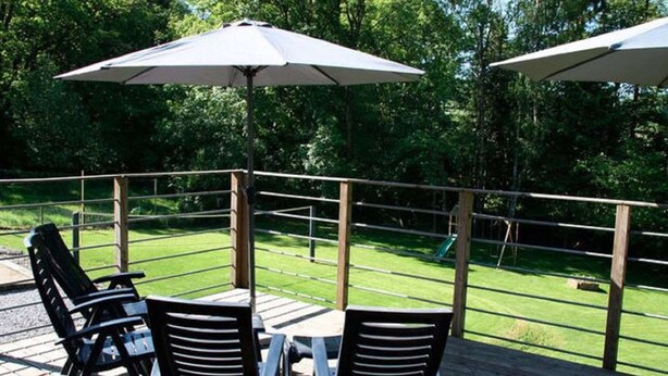 Una terraza exterior con mesa y sillas, rodeada por una barandilla de madera, con vistas a un área verde.
