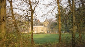 A large, historic stone building surrounded by a lush green field and dense trees. - Delightful Castle in Bastogne with Hot Tub (Bastogne)