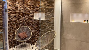A modern bathroom with a stone wall, two wicker chairs, a glass shower enclosure, and a shelf with toiletries. - Delightful Castle in Bastogne with Hot Tub (Bastogne)