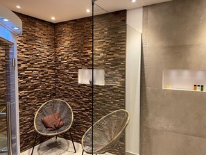 A modern bathroom with a stone wall, two wicker chairs, a glass shower enclosure, and a shelf with toiletries. - Delightful Castle in Bastogne with Hot Tub (Bastogne)