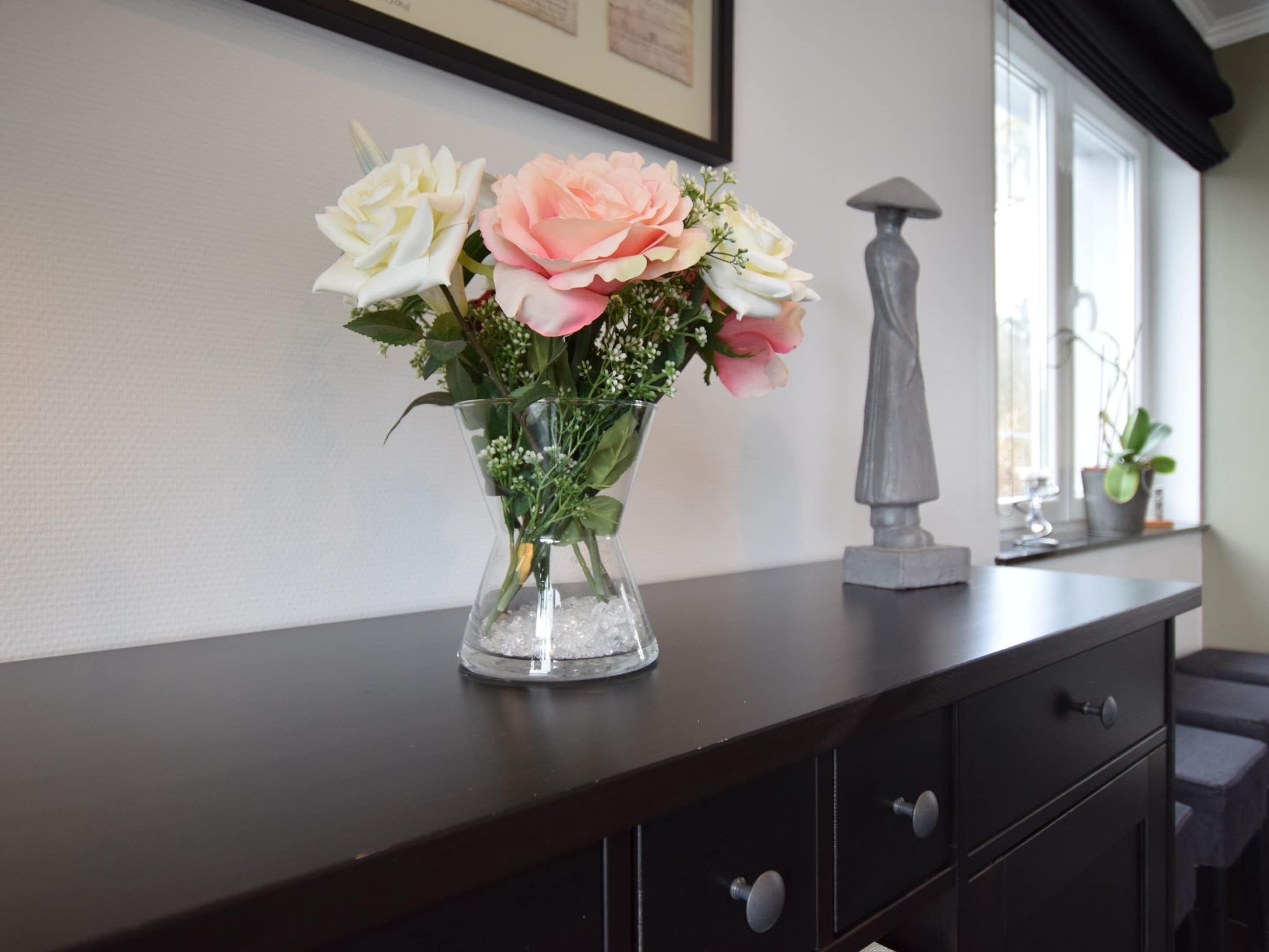 Un vase rempli de fleurs est posé sur une commode en bois sombre, dans une pièce dotée d’une fenêtre et d’une statue.