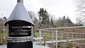 Un horno moderno para pizza al aire libre con una parrilla metálica, situado en una zona pavimentada cerca de una barandilla metálica y un pequeño puente sobre una fuente de agua.