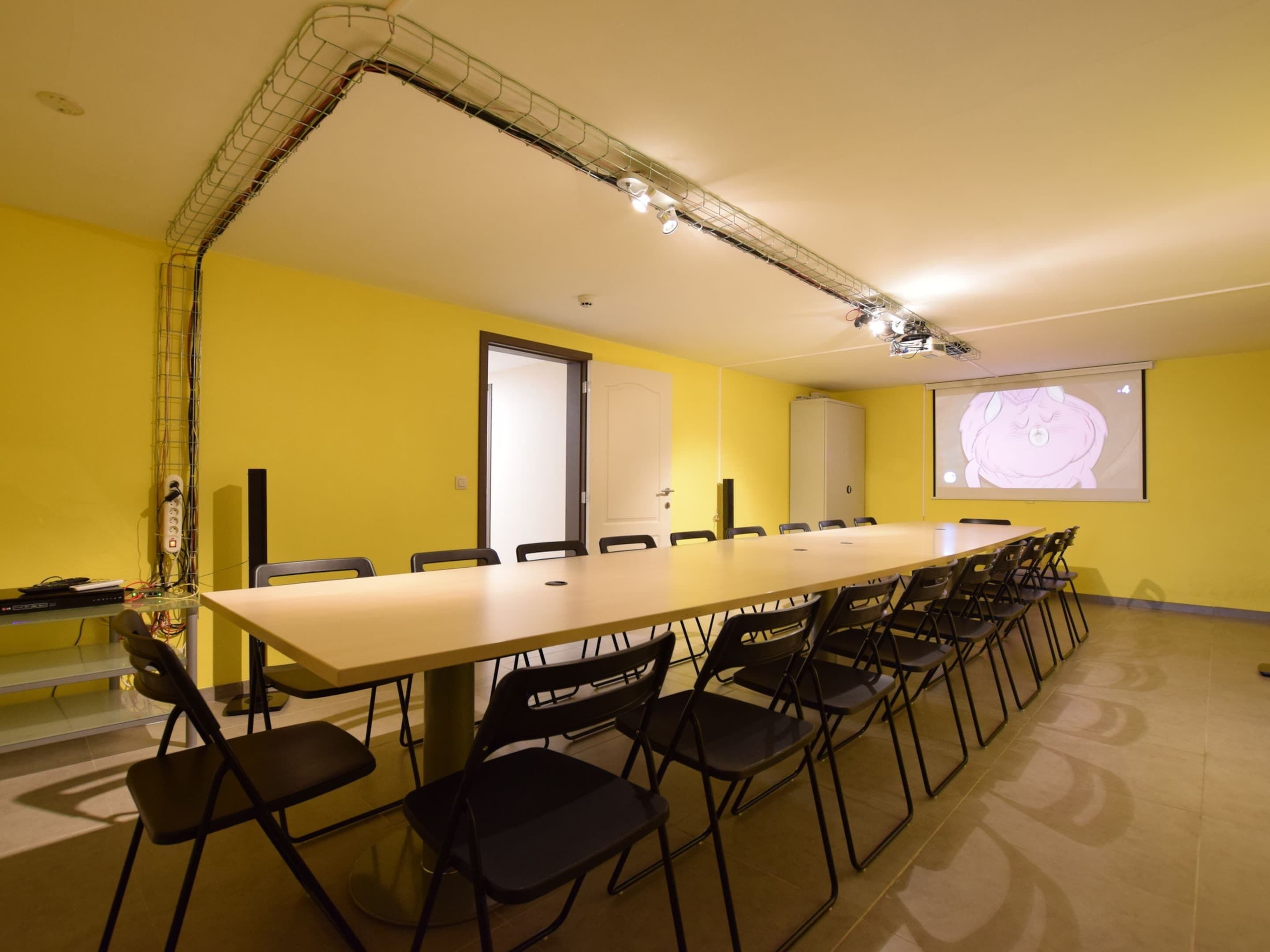 Un salón de conferencias con una larga mesa y sillas, una pantalla de proyector y una pared amarilla.