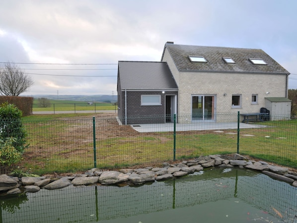 A modern two-story house with a sloped roof, a fenced yard, and a small pond.