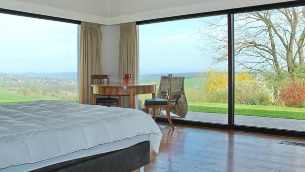 A bedroom with a large bed, a wooden table, a chair, and a view of a green landscape through large windows. - House in Somme-leuze Near Ardennes Hills (Somme-Leuze)