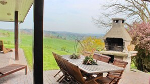 An outdoor patio with a stone barbecue grill, wooden table and chairs, and a view of rolling hills. - House in Somme-leuze Near Ardennes Hills (Somme-Leuze)