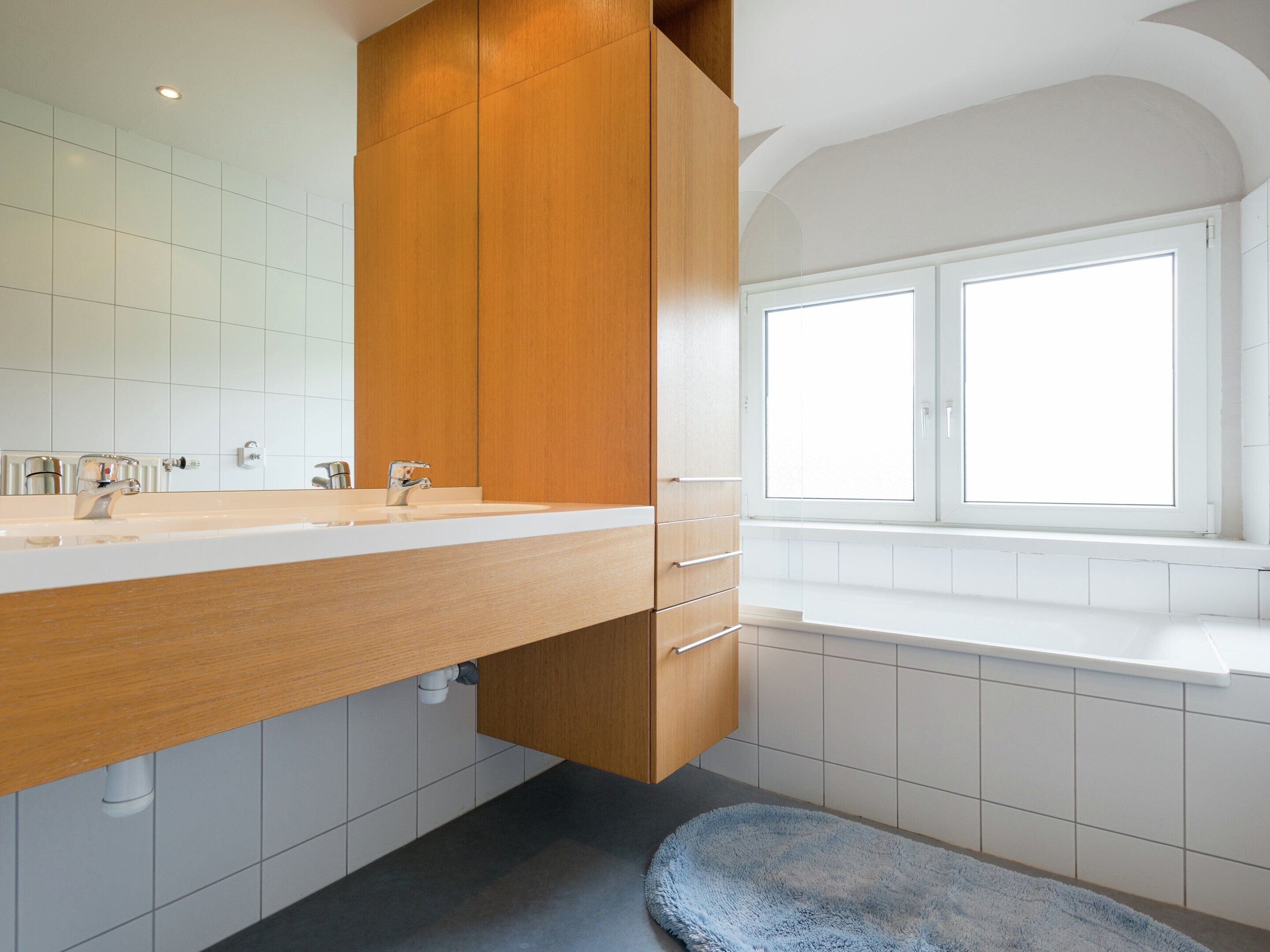 Une salle de bain moderne avec un meuble-lavabo en bois, un grand miroir, un lavabo blanc et une fenêtre au-dessus de la baignoire.