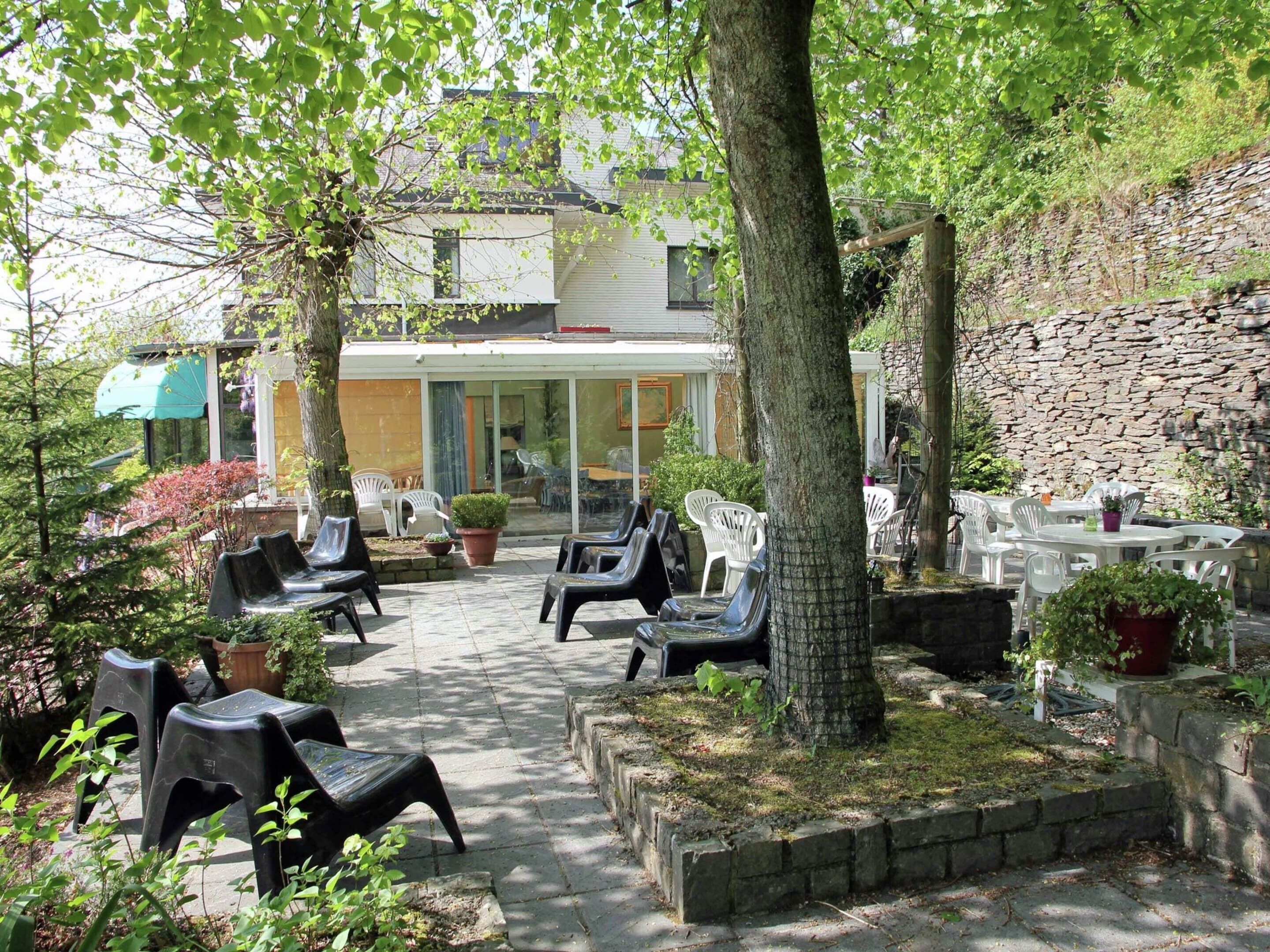Ein Terrassenbereich im Freien mit verschiedenen Sitzgelegenheiten, darunter Plastikstühle und Tische, umgeben von Grünpflanzen und einer Steinsäule.