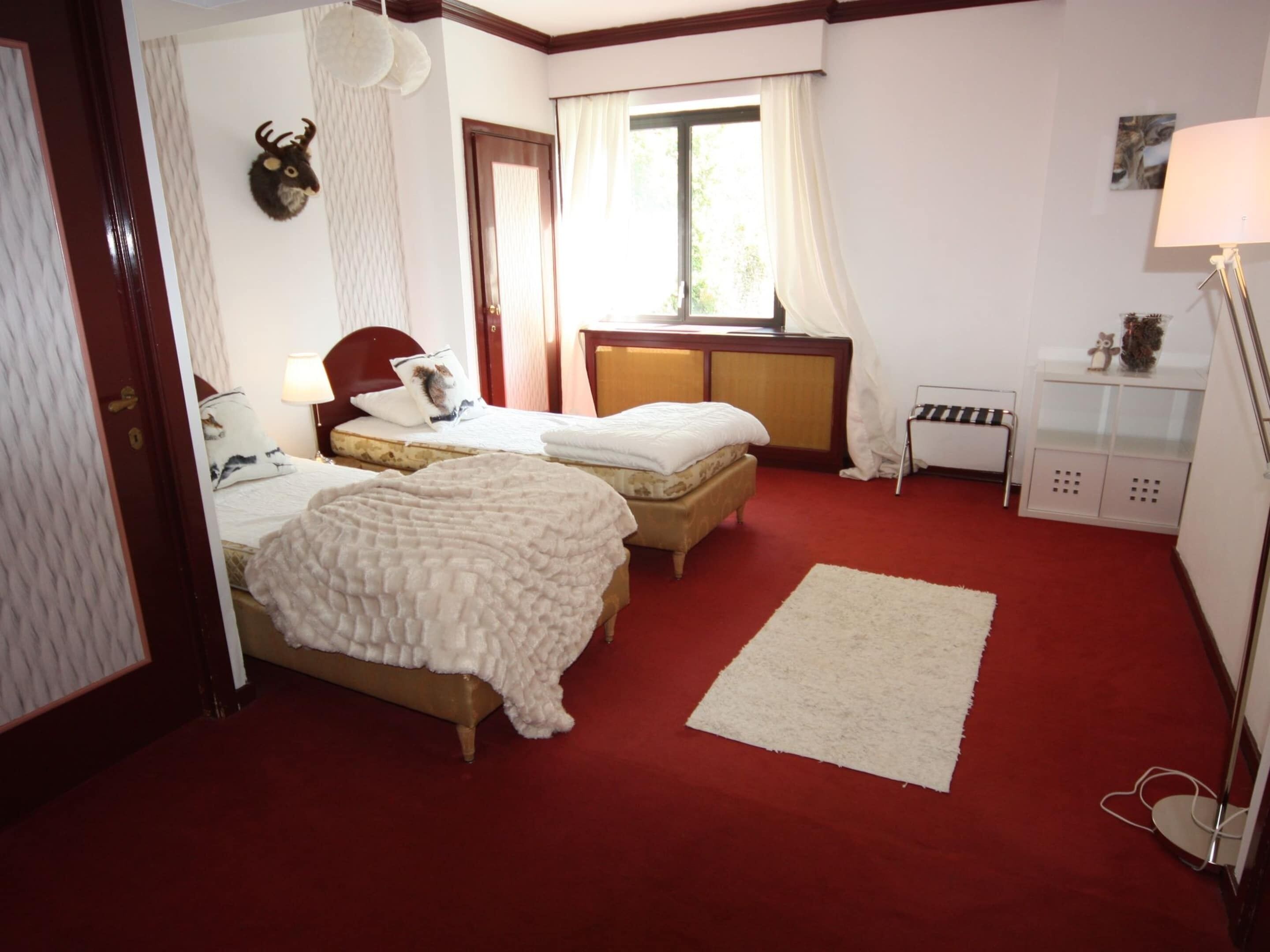 A hotel room with two beds, a red carpet, a white rug, a small table with a lamp, and a window with curtains.