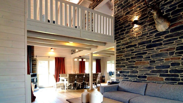 A spacious living area with a high ceiling, a stone wall, and a balcony with white railings. - Luxurious Villa with Sauna, Hot Tub, Recreation Room, Large Enclosed Garden (Durbuy)
