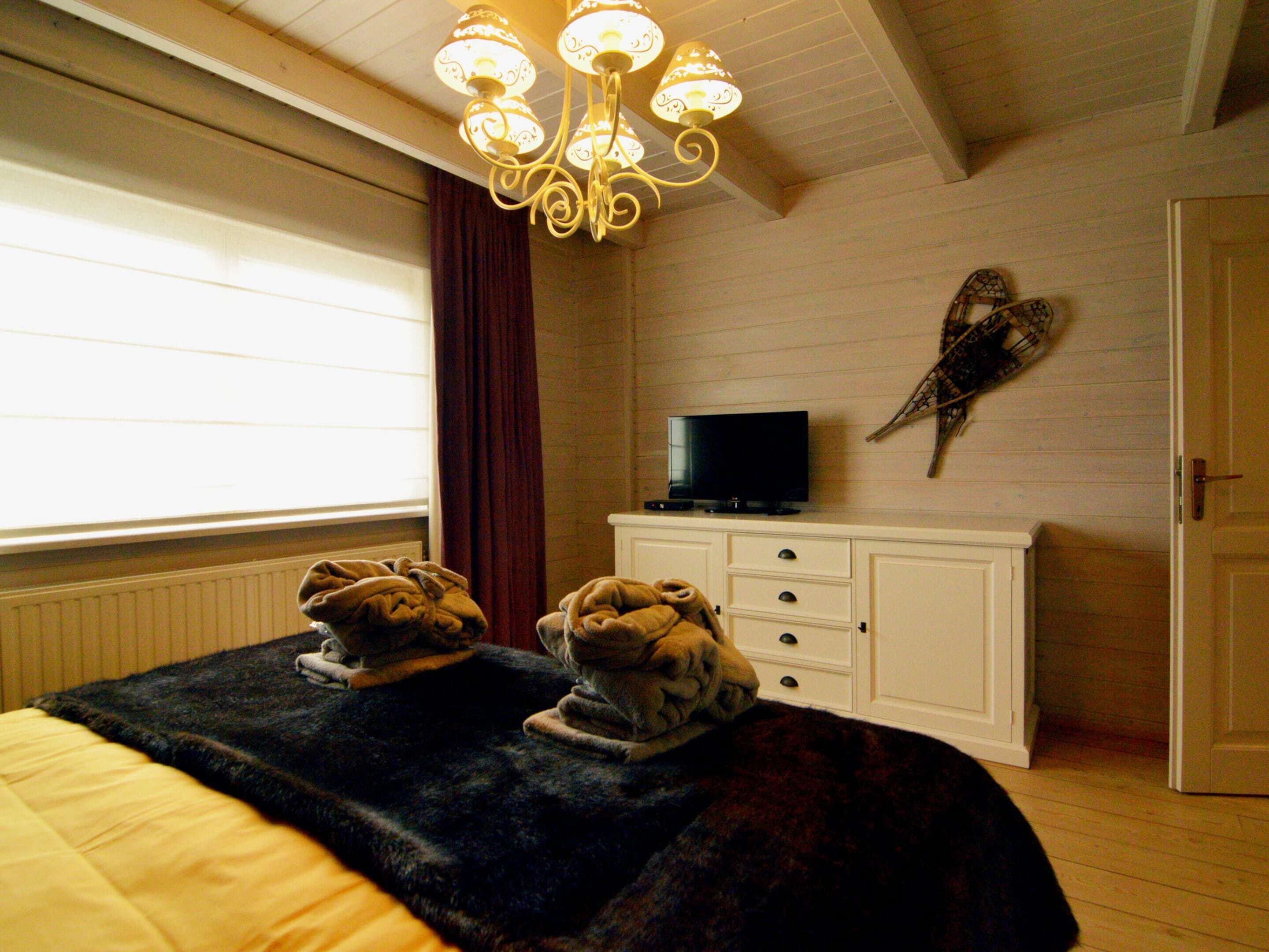 Ein Schlafzimmer mit einem Bett, einem Fernseher auf einer Kommode, einem Fenster mit Vorhängen und einer Pendelleuchte.