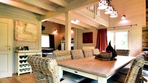 A dining area with a long wooden table surrounded by wicker chairs, a white cabinet with wine bottles, and a red curtain. - Luxurious Villa with Sauna, Hot Tub, Recreation Room, Large Enclosed Garden (Durbuy)