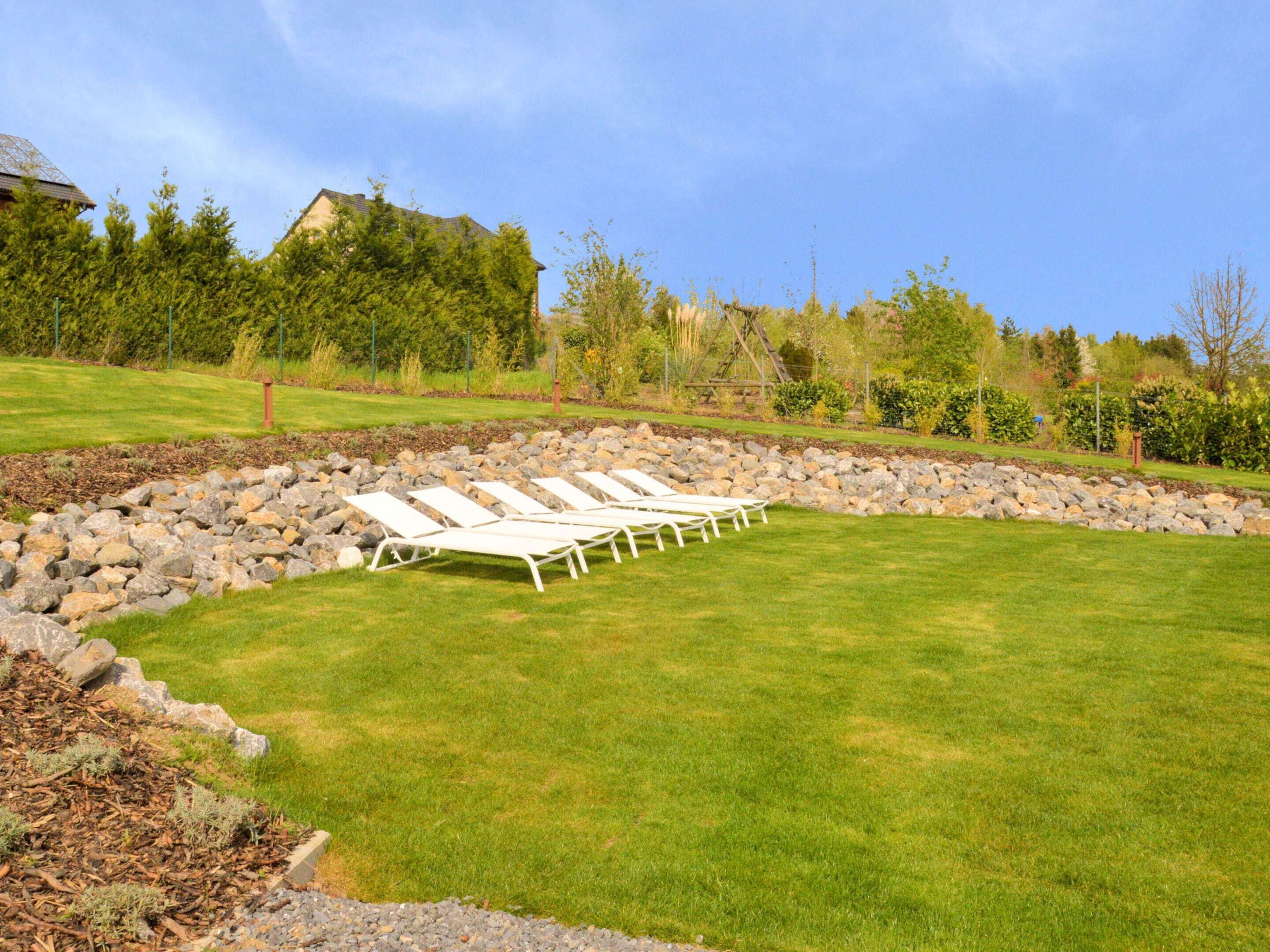 A well-maintained lawn with white lounge chairs arranged in a row, a stone retaining wall, and a variety of trees and shrubs in the background.